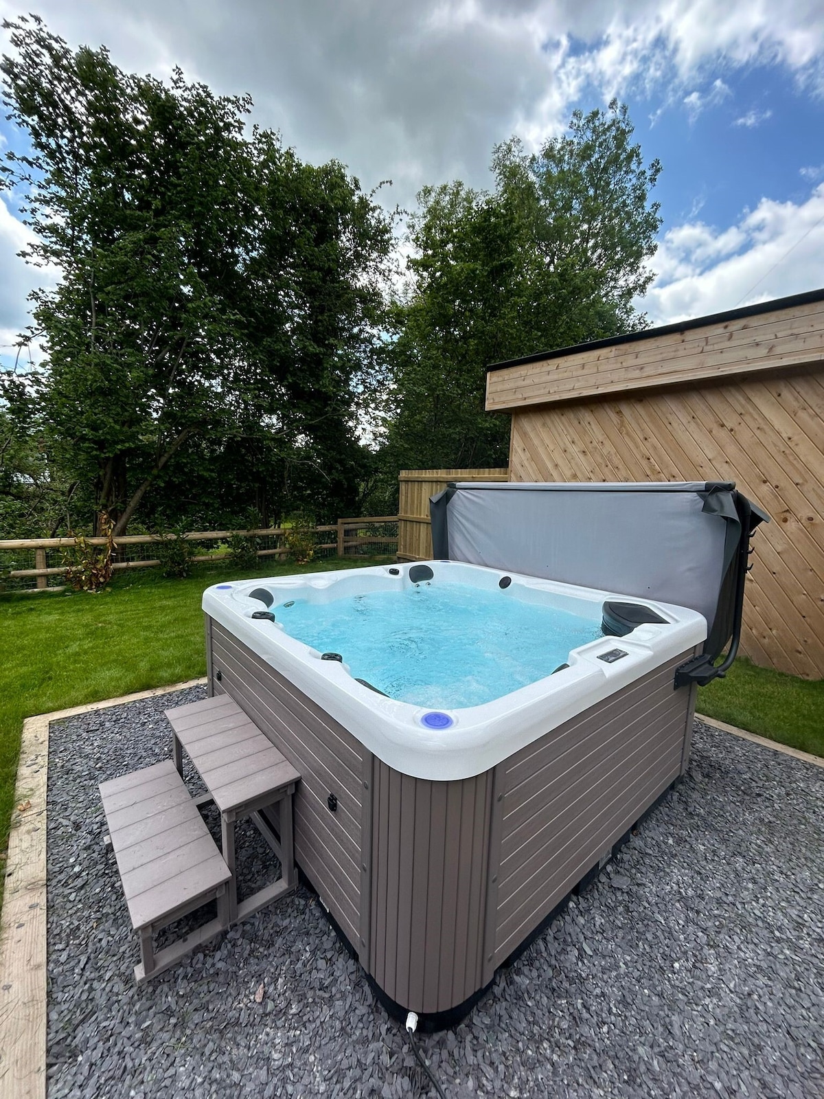 A hot tub sits on a stone-paved area surrounded by a lush green lawn. Wooden steps lead to the tub, which is covered by a grey protective cover. Tall trees and a wooden fence create a natural enclosure, enhancing the peaceful outdoor setting.