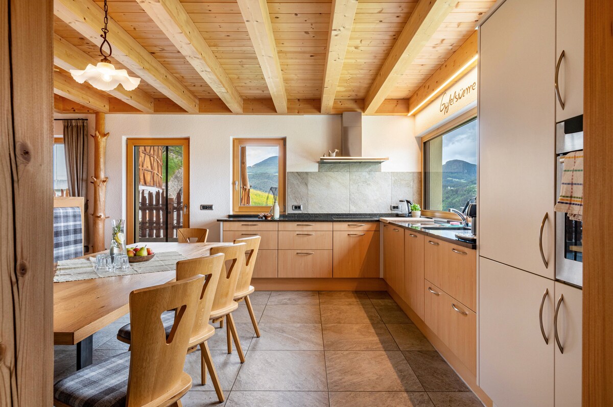 A modern kitchen is presented with warm wooden cabinetry and spacious countertops. A large wooden dining table accommodates six chairs, enhancing the communal atmosphere. Large windows allow natural light to illuminate the space, offering views of the surrounding landscape.