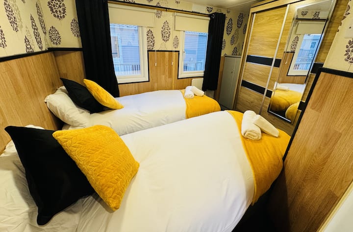 חדר שינה זוגי מואר עם מיטות נוחות, כריות נוי צהובות וארון בגדים עם מראה למרחב נוסף. מושלם לשהייה רגועה עם עיצוב מסוגנן ואור טבעי.