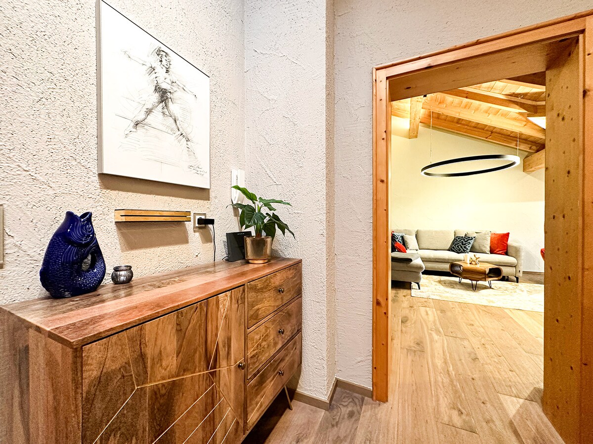 A wooden sideboard with a geometric design sits against the wall. A small plant and decorative items are displayed on top. A doorframe leads into a spacious living area with a cozy sofa and a circular light fixture overhead.