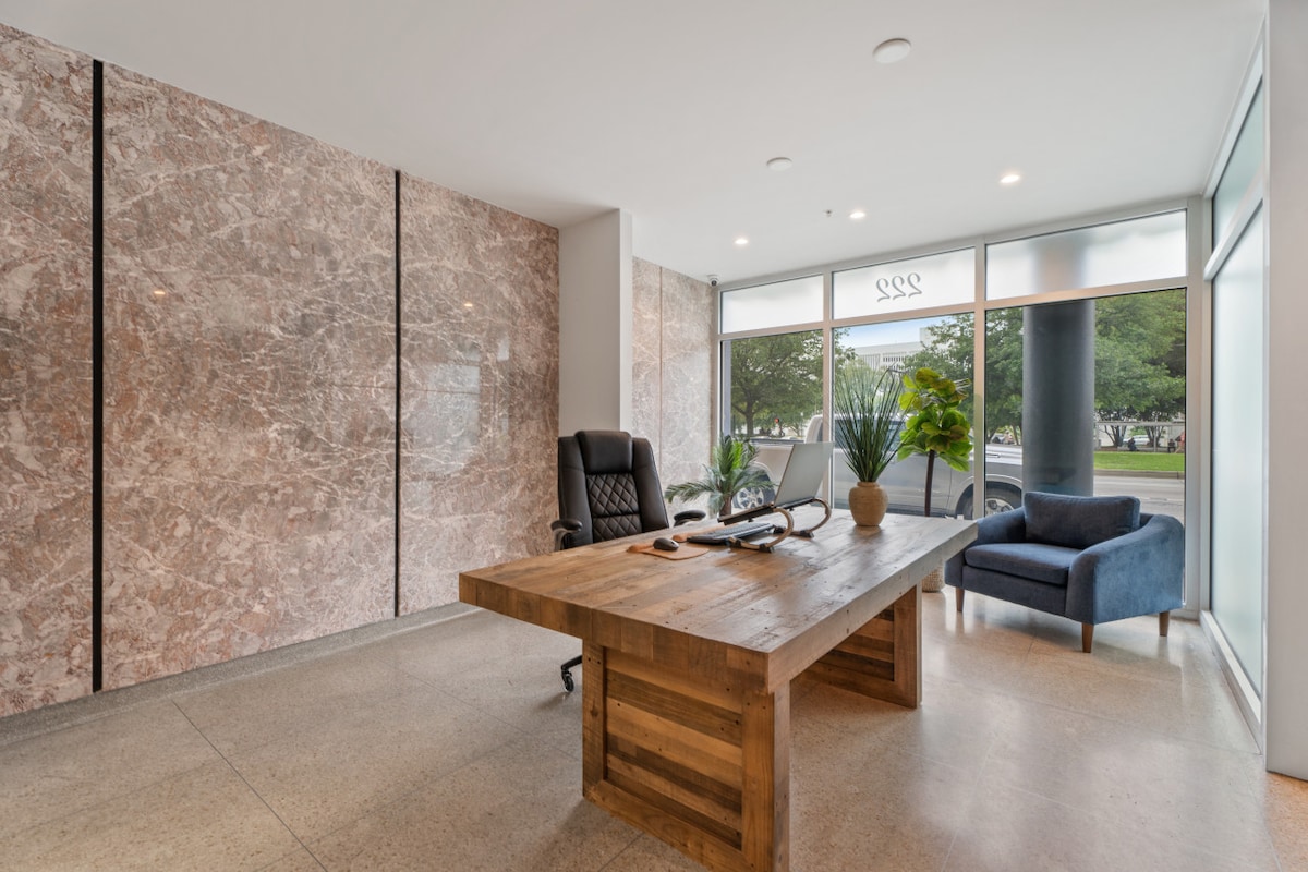 A sleek reception area is presented with a large wooden desk and a comfortable blue armchair. The room features smooth marble walls and large windows that allow natural light to illuminate the space, creating an open and inviting atmosphere.