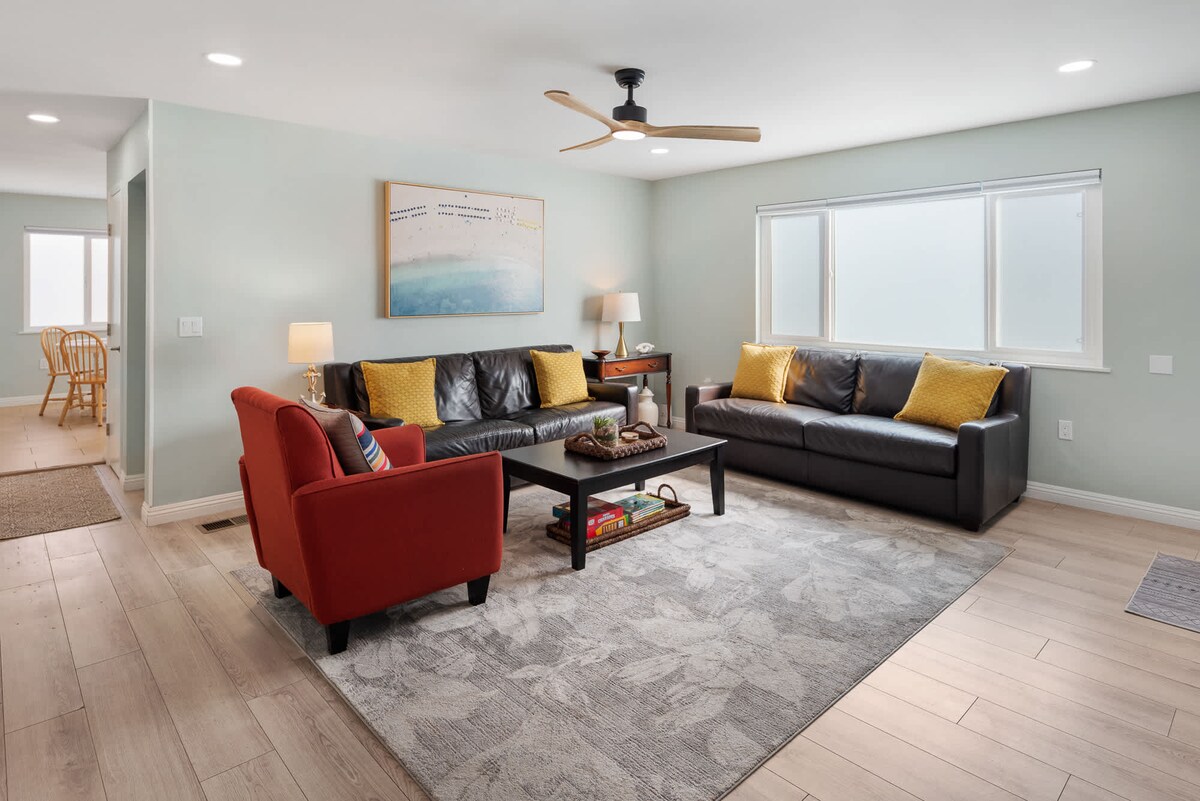 The living area features two comfortable sofas with bright yellow accent pillows and a vibrant red armchair. A coffee table sits in the center on a muted area rug. Natural light fills the space through large windows, highlighting the soothing wall color.