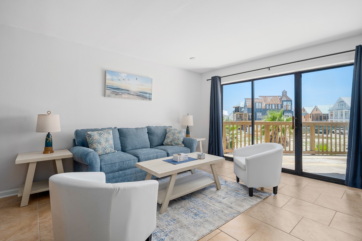 The living area features a comfortable blue sofa with coordinating cushions, complemented by two white armchairs. A light wooden coffee table sits atop a rug, while natural light floods the space through expansive glass doors that lead to a private balcony.