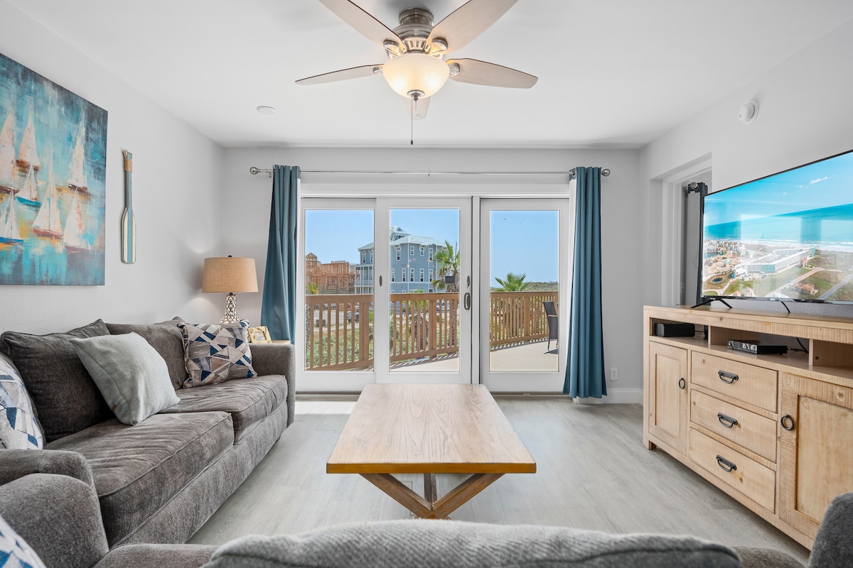 A comfortable living area features a large flat screen TV and a spacious gray sofa adorned with decorative pillows. Natural light enters through sliding glass doors, leading to a private patio. A wooden coffee table centers the arrangement, contributing to the inviting atmosphere.