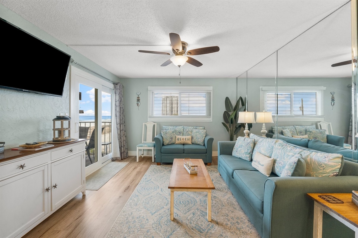 A light-filled living area features a ceiling fan and large mirrors enhancing the sense of space. A comfortable seating arrangement includes turquoise sofas adorned with decorative pillows. A coffee table rests in the center, while sliding glass doors lead to a balcony with ocean views.