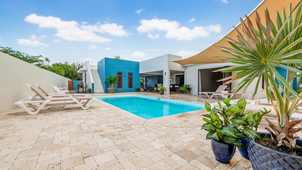A private pool area is surrounded by natural stone tiles, featuring a refreshing swimming pool illuminated by sunlight. Comfortable loungers are positioned nearby, accompanied by potted plants and vibrant greenery, providing a relaxing outdoor environment suitable for leisure and sunbathing.