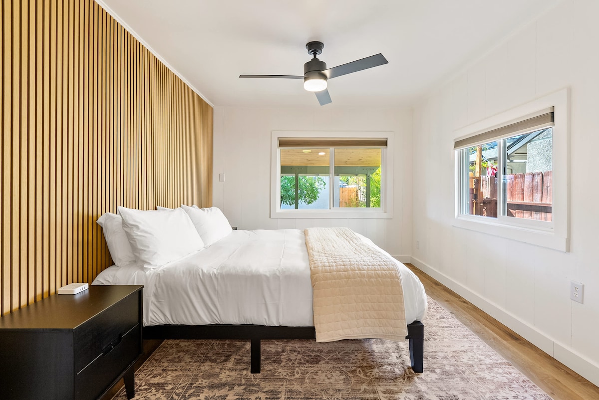 A spacious bedroom features a large bed with crisp white linens and a decorative throw. The room is illuminated by natural light from two windows, with a modern ceiling fan overhead and warm wood accents on the walls.