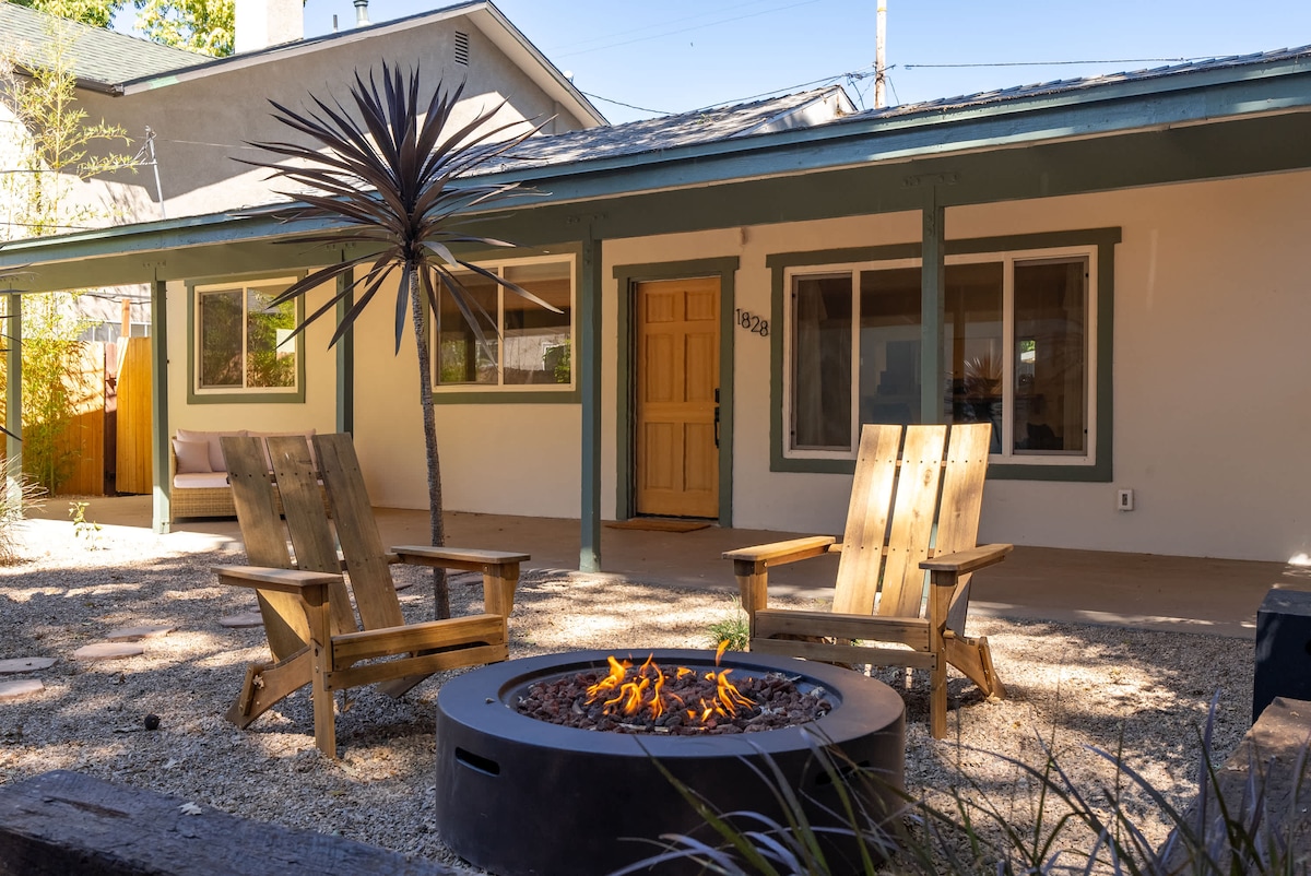 An outdoor seating area features two wooden chairs arranged around a circular firepit, creating a cozy atmosphere. The background includes a single-story home with large windows, and a pathway leads to the front door. Landscaping adds natural beauty to the private space.
