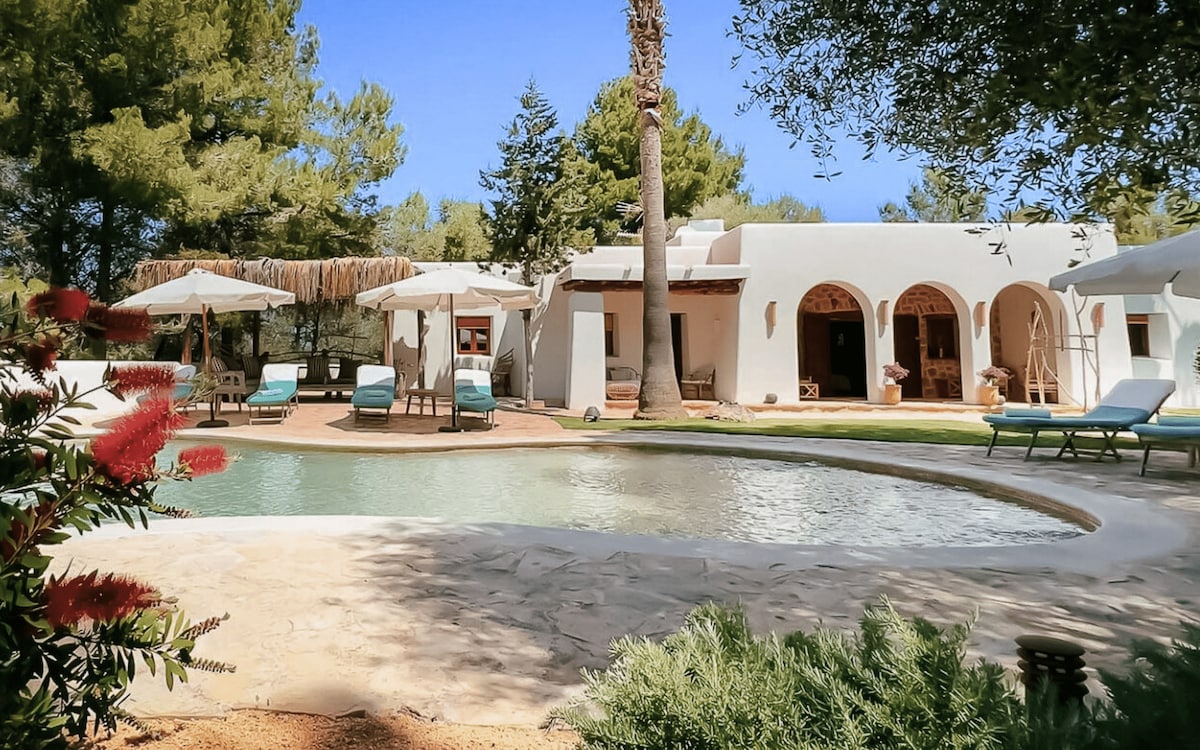 A serene outdoor area features a gentle saltwater pool surrounded by lush greenery. Sun loungers are positioned under sun umbrellas, providing comfort for relaxation. The charming architecture of the retreat complements the natural scenery, offering a peaceful ambiance.