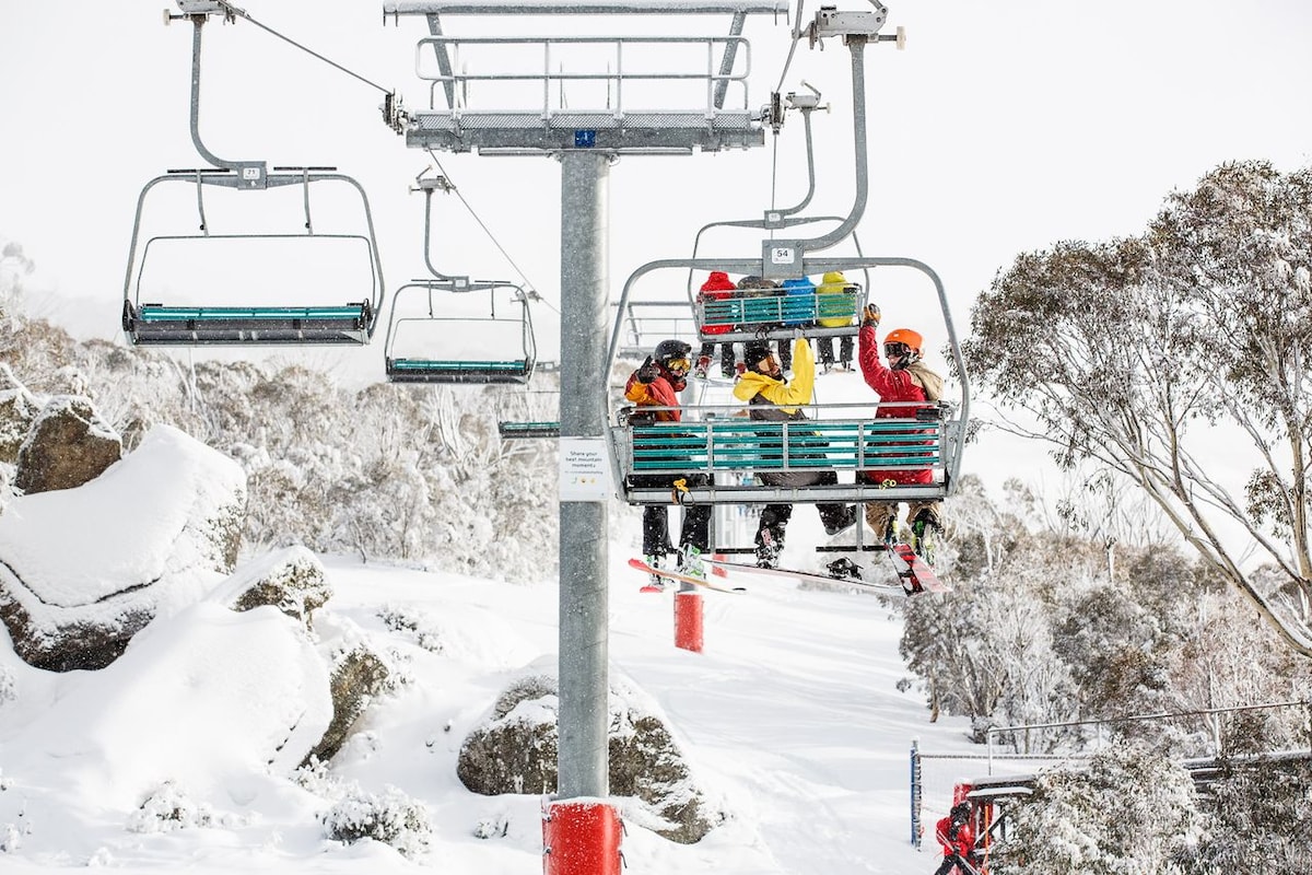 A ski lift transports riders up a snow-covered slope, surrounded by frosty trees and rocky outcrops. The chairs are filled with individuals dressed in colorful winter gear, ready to enjoy the snowy landscape. Snow blankets the ground, creating a serene winter atmosphere.