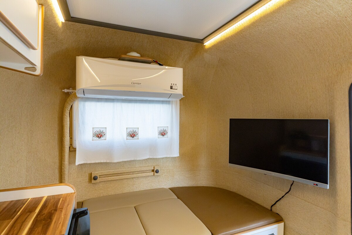 A compact interior showcases a cozy sleeping area with a plush double bed. Above, a flat-screen TV is mounted on a light-colored wall. Soft lighting highlights the warm tones of the space, complemented by a window adorned with a simple curtain.
