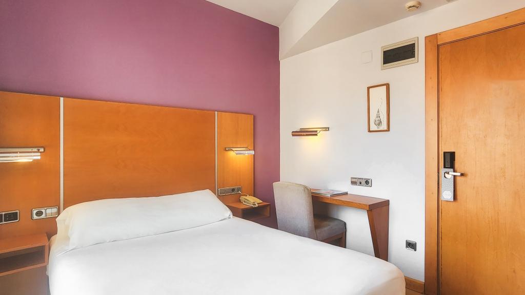 A cozy sleeping area is designed with a light-colored bedspread atop a spacious bed. A contrasting purple wall enhances the ambiance. A wooden desk and chair are positioned beside the bed, accompanied by functional wall-mounted lights.
