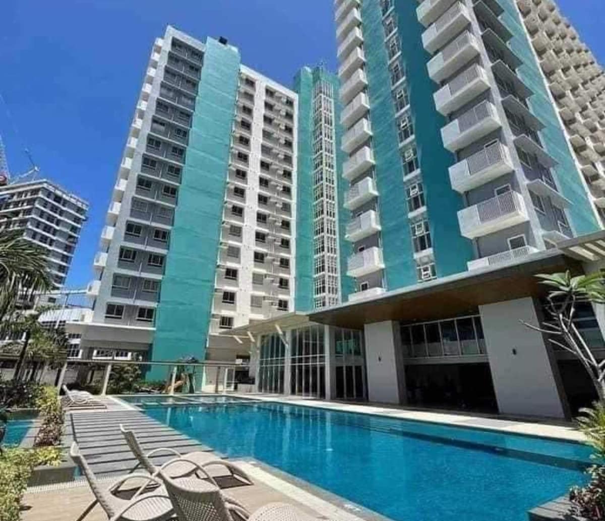 A modern building complex is displayed with two tall structures featuring vibrant turquoise exteriors. A spacious outdoor pool is located in the foreground, surrounded by loungers and tropical plants, creating an inviting atmosphere in the sunny environment.
