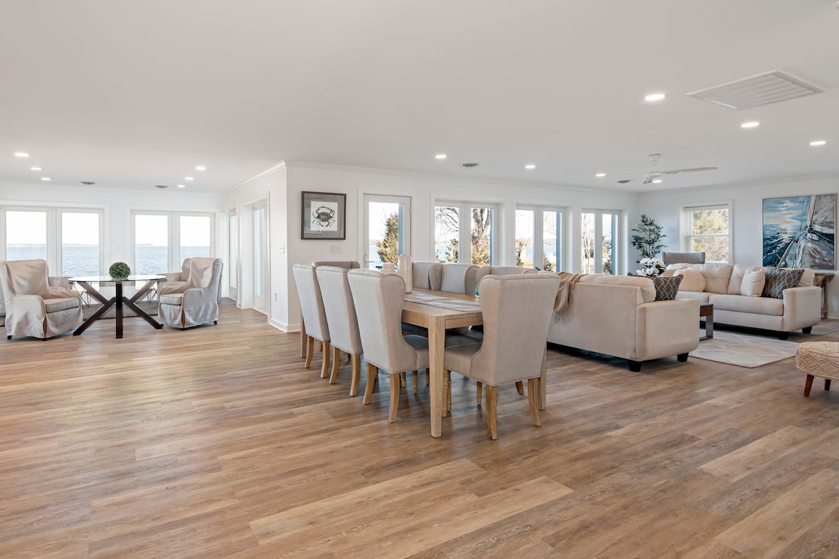 A spacious open-concept living area features large windows allowing natural light to illuminate the room. Light wood flooring enhances the inviting atmosphere. Comfortable seating arrangements are provided, including a sectional sofa and a large dining table surrounded by fabric chairs, all set against a backdrop of water views.