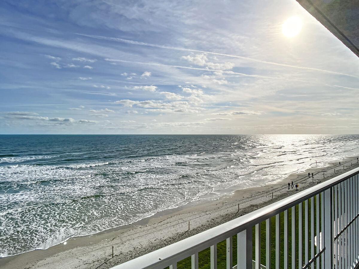 An expansive ocean view is captured from the private balcony, showcasing a shimmering shoreline under bright sunlight. Gentle waves roll onto the sandy beach, while people can be seen walking along the water's edge, highlighting the tranquil atmosphere of the seaside.