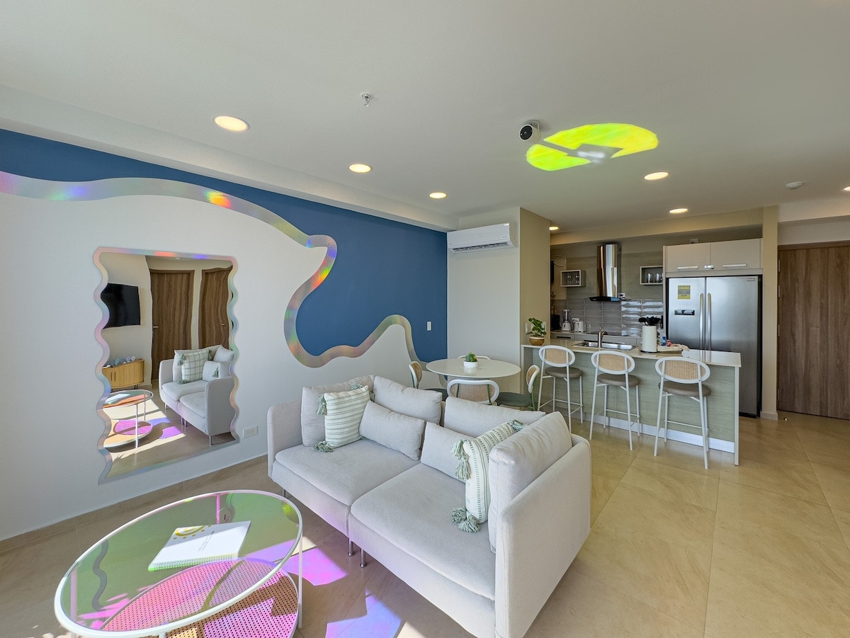 A modern living space is showcased, featuring a light-colored sofa accompanied by a glass coffee table. A vibrant wall mural adds a splash of color. The kitchen area is equipped with stainless steel appliances, and a large mirror reflects the inviting layout.