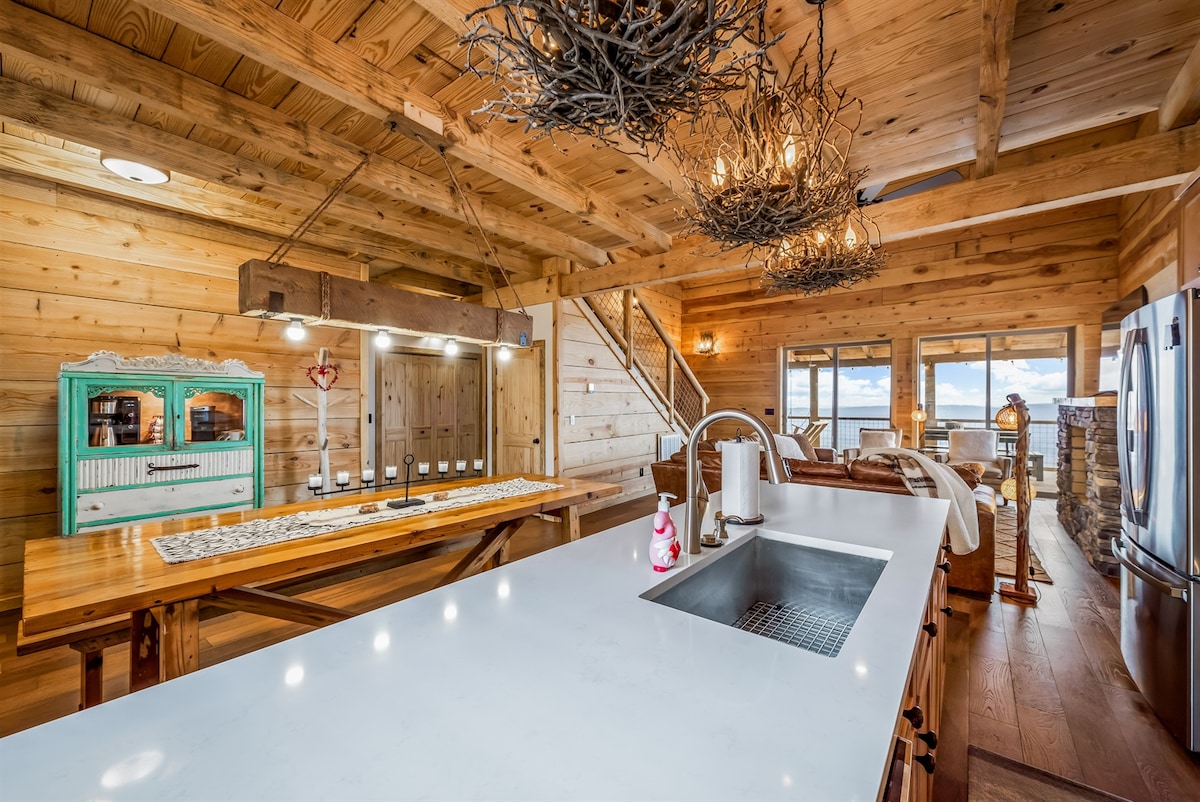 A spacious kitchen and dining area is presented with a large wooden dining table and a well-equipped counter with a sink. Natural wood finishes and unique light fixtures add character, while large windows offer views of the outdoors, inviting ample natural light into the room.