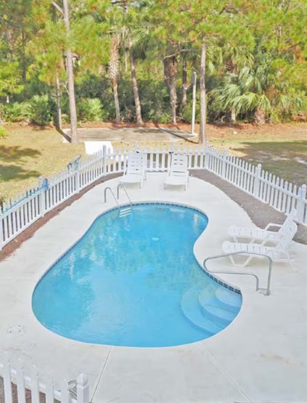 A private swimming pool is surrounded by a white picket fence, offering a serene outdoor space. Several reclining chairs are positioned around the pool, amidst lush greenery and tall trees. The inviting pool features a gradual entry and a smooth, curvy design.
