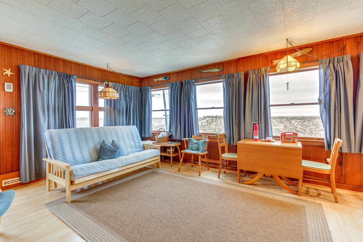 The living room features large windows draped with blue curtains, allowing natural light to filter in. A full futon is positioned alongside a dining table and chairs, while wood paneling adds a warm touch. A textured area rug lies on the floor, enhancing the cozy atmosphere.