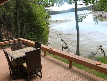 A spacious wooden deck features a dining table with four chairs, positioned to overlook the water's edge. Surrounding trees provide a natural setting, with views of the water, which appears calm and reflective.