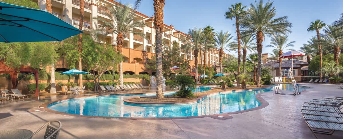 A spacious outdoor pool area is surrounded by lush palm trees and resort buildings. Umbrellas provide shade by the water's edge, and lounge chairs are arranged along the poolside. The pool's curved design complements the relaxing atmosphere.