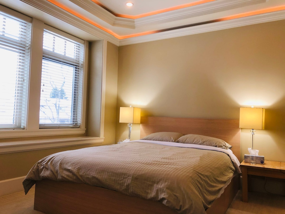 A spacious bedroom is featured with a large bed dressed in neutral tones. Two bedside lamps provide soft lighting, and natural light filters through the window with blinds. The room is finished with an elegant ceiling design and warm hues along the edges.