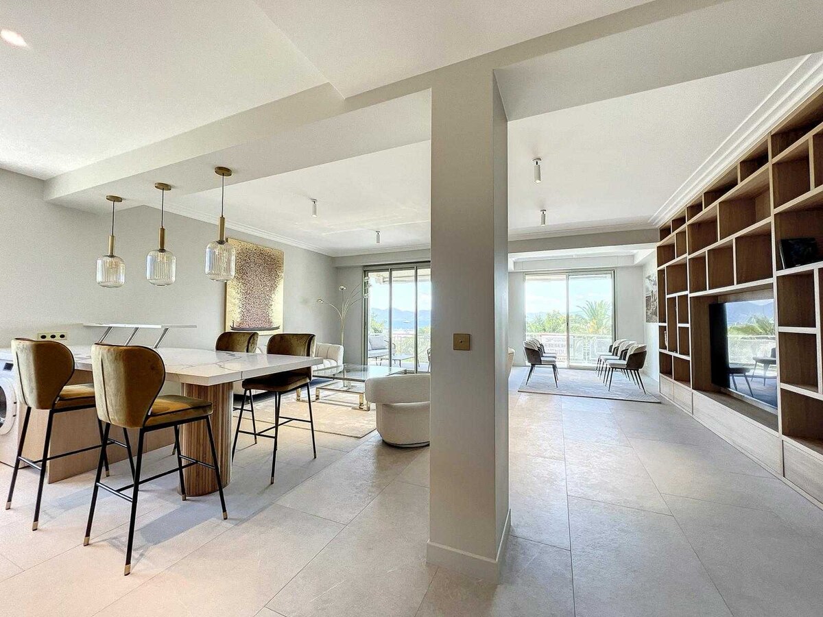 An open living space is captured, featuring a modern dining area with a large wooden table and stylish bar stools. Natural light fills the room, highlighting a comfortable seating area and an elegant wall unit. Expansive glass doors lead to a terrace with outdoor views.