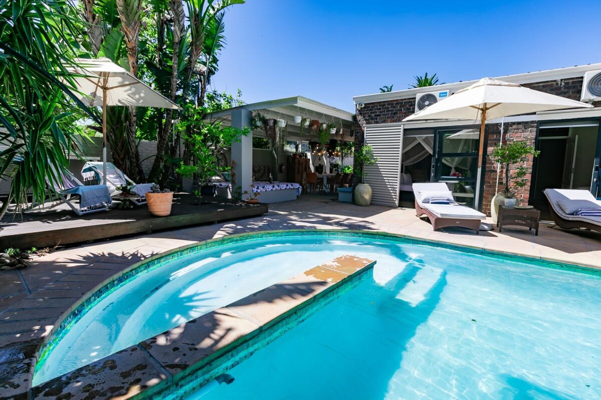 A refreshing pool is surrounded by lush greenery, featuring sun-beds and parasols for relaxation. An outdoor dining area is visible in the background, offering a shaded space for meals. The space is well-lit, creating an inviting atmosphere for outdoor gatherings.