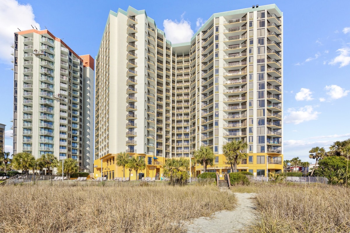 A modern beachfront building stands tall against a clear sky, with multiple floors featuring balconies. Lush palm trees and dry grass are visible in the foreground, leading the way toward the entrance of the resort, which is brightly painted in warm tones.