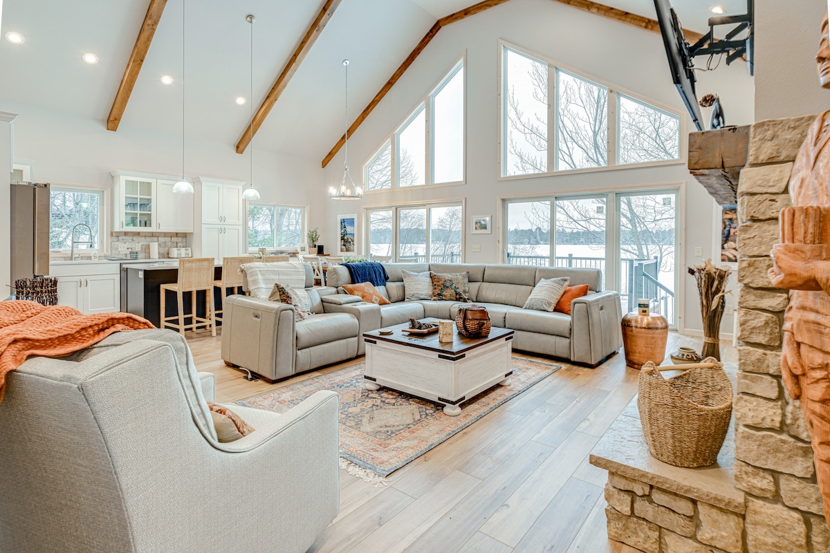 The spacious living area features high vaulted ceilings with exposed wooden beams. Large windows provide abundant natural light and showcase lake views. A comfortable sectional sofa encircles a decorative coffee table, with additional seating available in an accent chair. A stone fireplace adds warmth to the room.