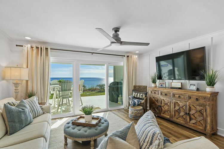A light-filled living area is presented, featuring a comfortable white sofa with blue and white accents. A large TV is mounted on the wall across from a woven chair, while sliding glass doors offer views of the Gulf. Elegant curtains frame the windows, enhancing the coastal feel.