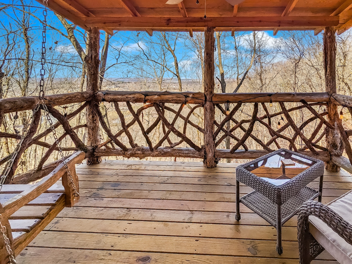 A wooden porch offers a relaxed seating area with rustic log railings and a woven coffee table. A swing chair is positioned beside the railing, showcasing expansive views of the surrounding landscape with bare trees under a clear blue sky.