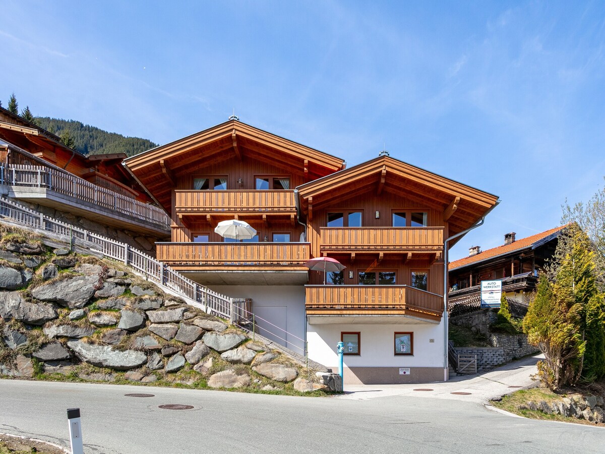 The chalet features a traditional wooden exterior with a sloped roof and spacious balconies. Natural stone steps lead up to the entrance, surrounded by landscaped greenery. Large windows reflect the mountainous landscape, providing a warm, inviting appearance.
