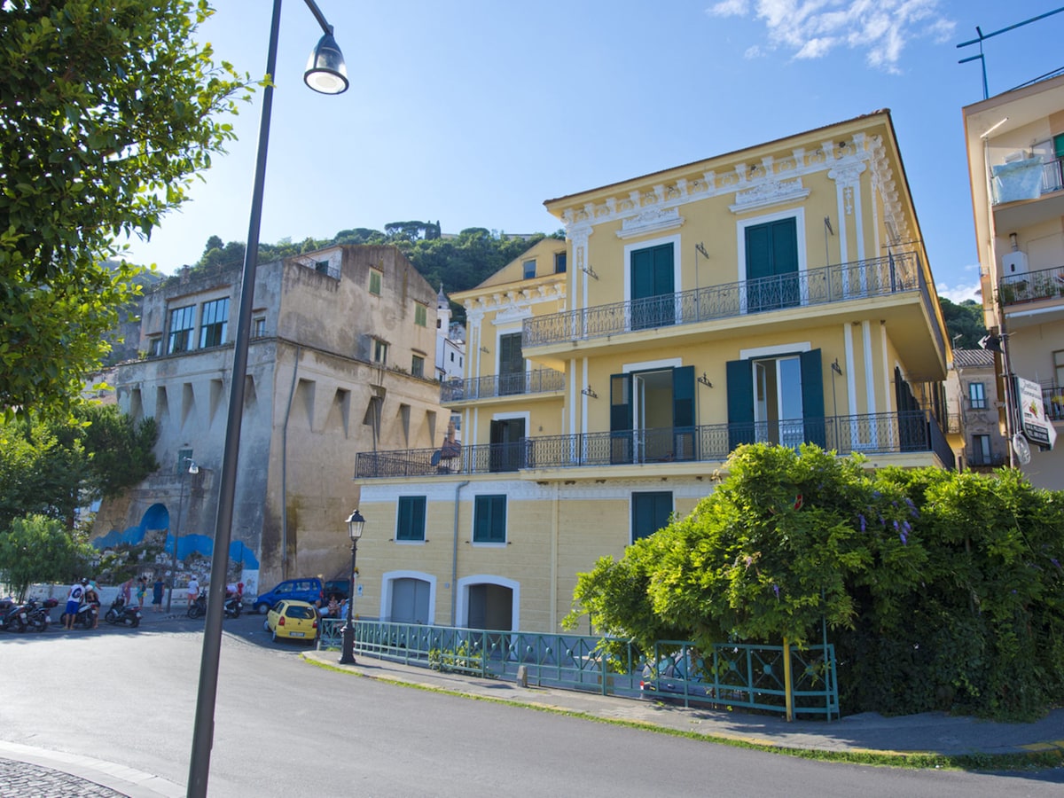 The image showcases a three-story yellow building with multiple balconies and large windows. Green shutters adorn the windows, and lush greenery is visible at the base. Nearby, a tree and street lamp are included, enhancing the urban landscape. A blue sky and nearby hill provide a scenic backdrop.