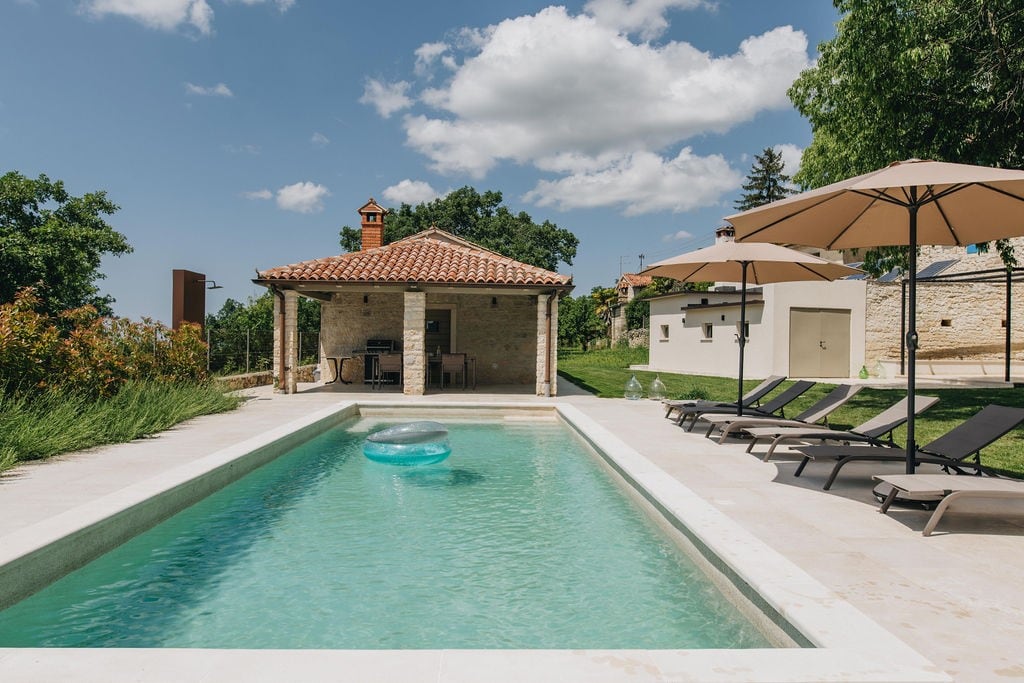 A tranquil pool area features a rectangular pool with a hydro-massage bench, surrounded by several deckchairs and large umbrellas. A charming stone building, with a terracotta roof, stands nearby, complementing the lush green landscape.