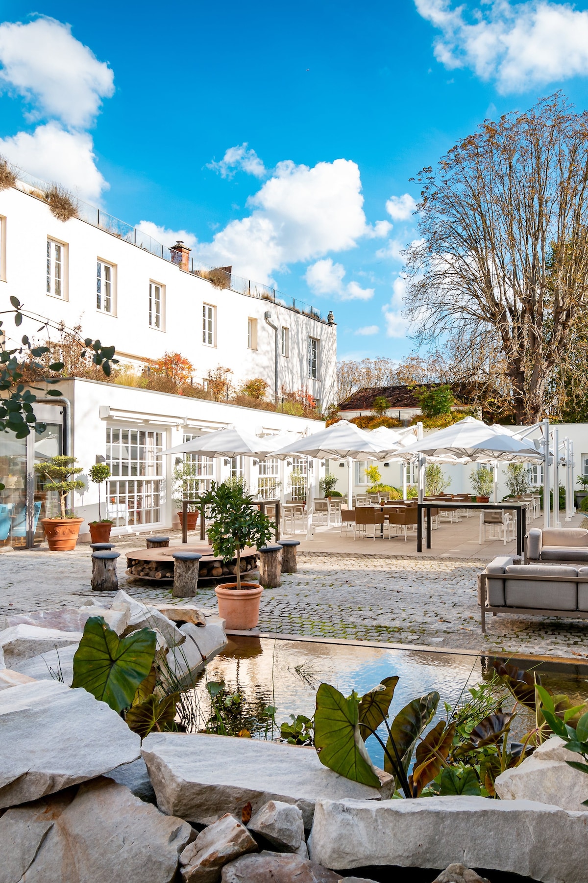 An outdoor terrace is framed by lush greenery and white structures, featuring an arrangement of seating and dining areas. Natural light illuminates the setting, while a small water feature adds a tranquil touch, creating an inviting space for relaxation.