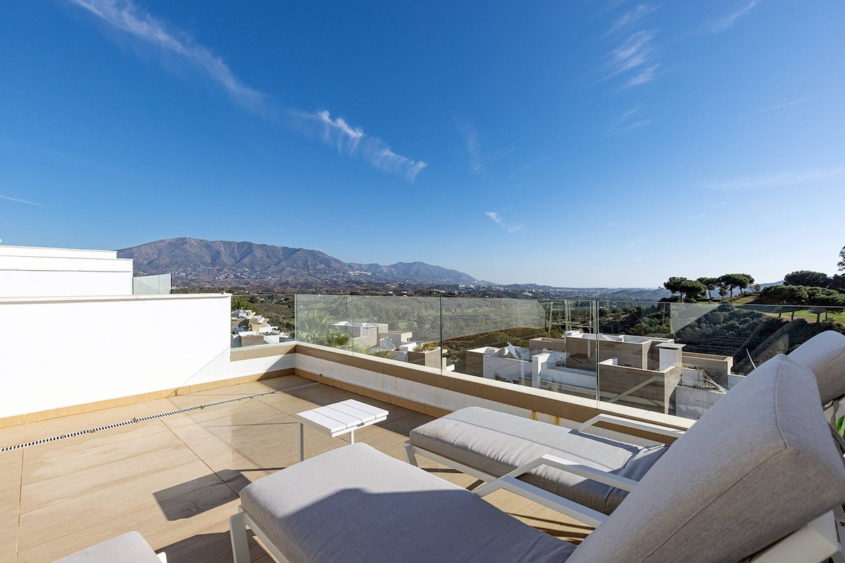 The rooftop terrace features sun loungers and a small dining table, set against a backdrop of expansive mountain views. The space is designed for relaxation, with clear glass railings enhancing the open feeling of the area, allowing for unobstructed views of the surrounding landscape.