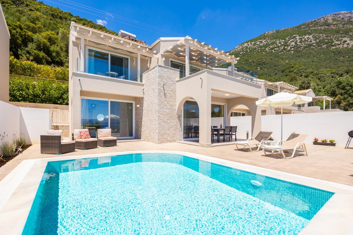 A modern villa exterior features a private pool surrounded by a furnished patio. Comfortable seating arrangements and sun loungers offer relaxation options. Large glass doors provide access to indoor living spaces, with the backdrop of a lush green hillside enhancing the serene atmosphere.