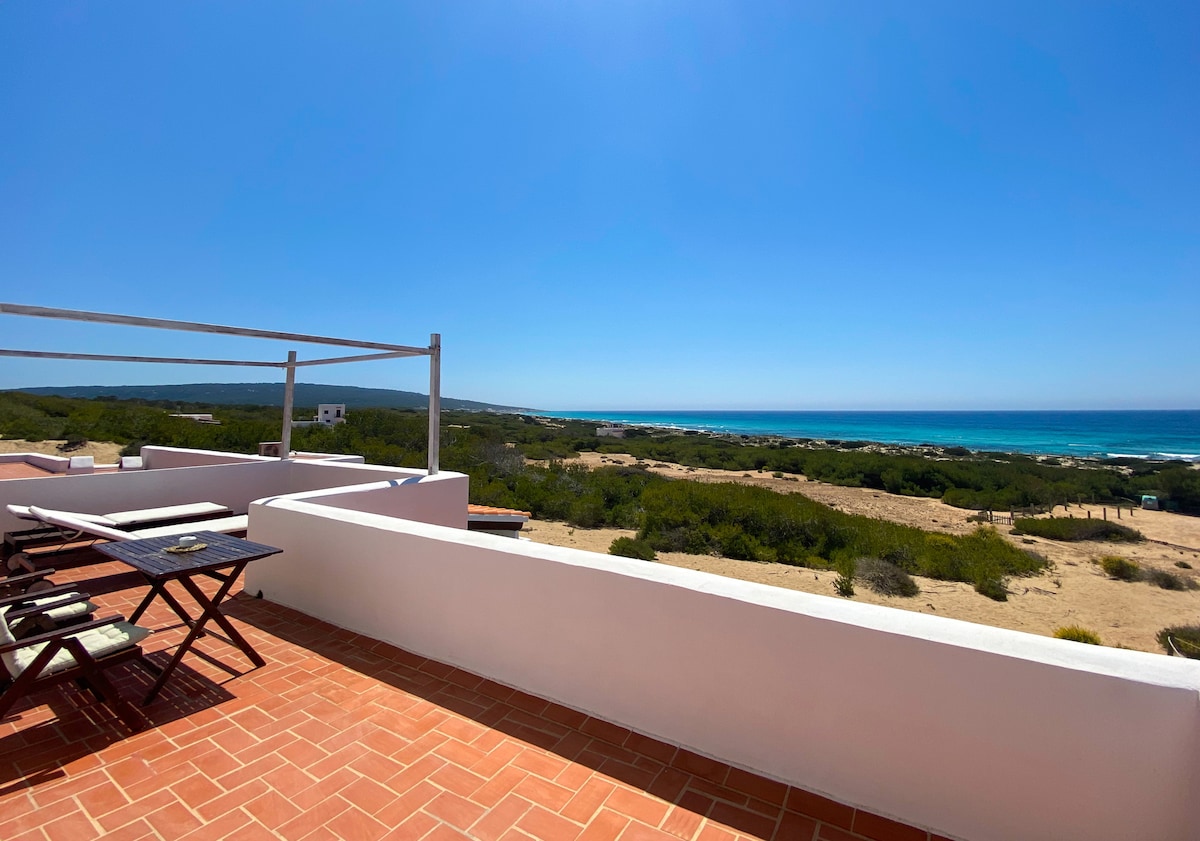 A spacious terrace offers panoramic sea views, with a wooden dining table and chairs positioned for outdoor dining. The surrounding landscape features greenery and distant shoreline, complemented by clear blue skies.