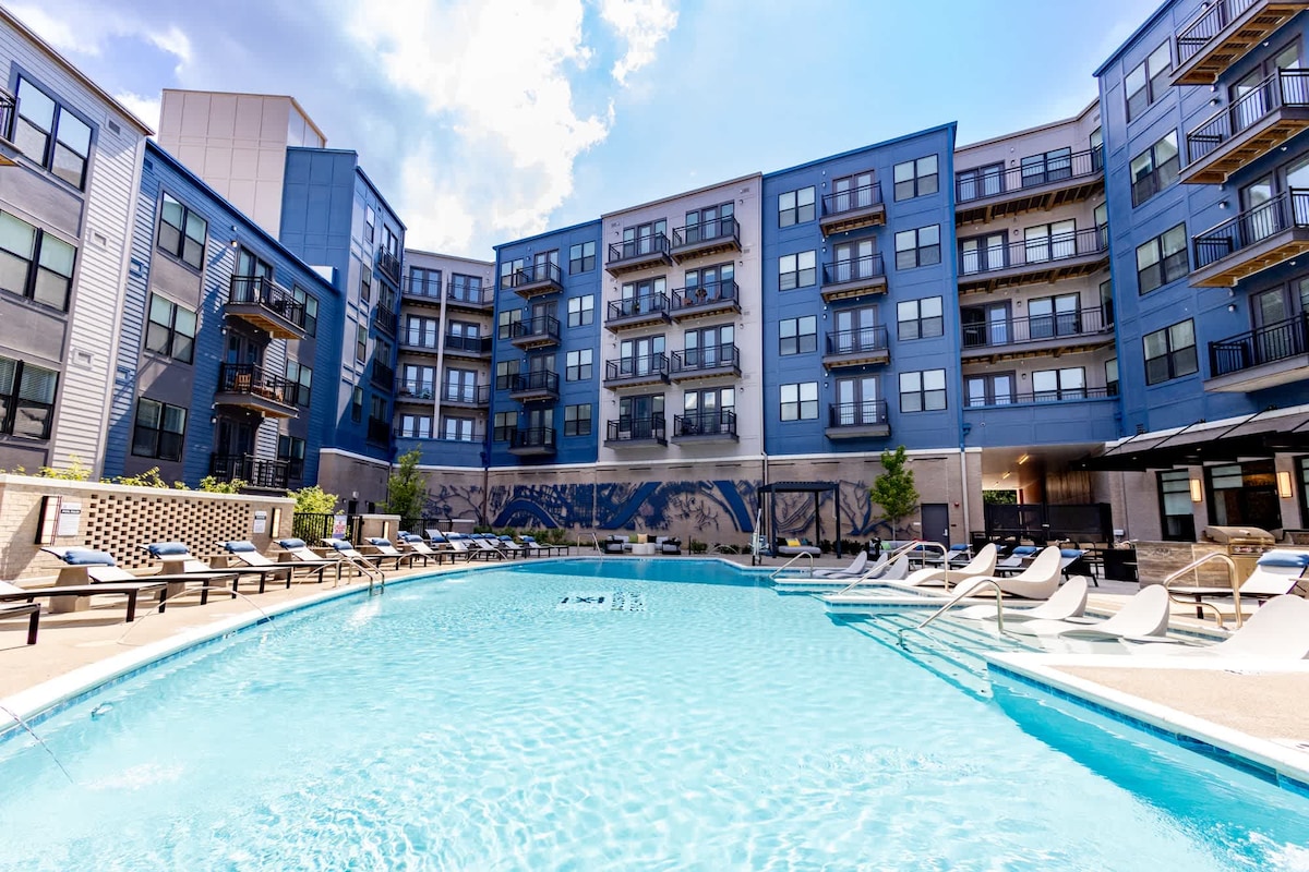 A modern pool area is showcased, featuring a rectangular swimming pool surrounded by lounge chairs. The multi-story building in the background has a contemporary design with large windows and balconies, offering a glimpse of the vibrant atmosphere. Lush greenery is visible along the edges.