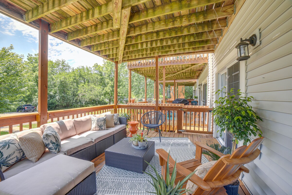 A spacious deck features comfortable seating arrangements, including a sectional sofa and wooden chairs. A coffee table sits centrally on a patterned rug, and greenery surrounds the area, creating a peaceful atmosphere for relaxation. An outdoor gas grill is visible in the background.