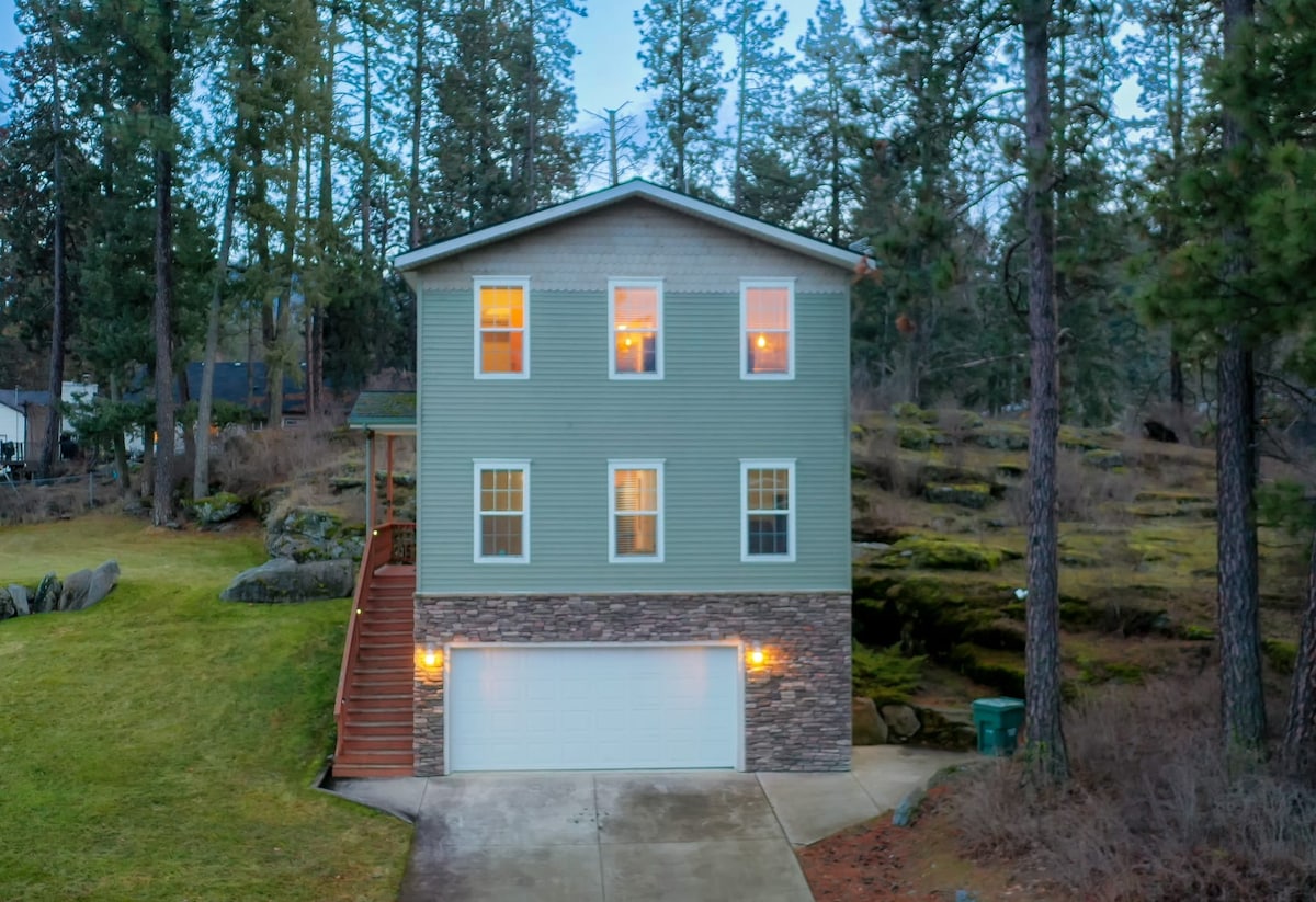 The exterior of a two-story home is seen, featuring a mix of siding and stone accents. Large windows allow warm light to glow from within. The house is surrounded by trees and natural rock formations, with a driveway leading to a garage.