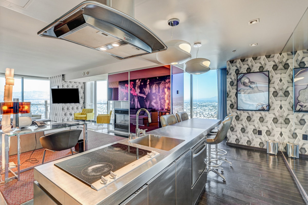An open-concept kitchen and living area is showcased, featuring modern stainless steel appliances and a spacious island with seating. Large windows offer expansive views of the surroundings, while vibrant art pieces and stylish furnishings create a contemporary atmosphere.