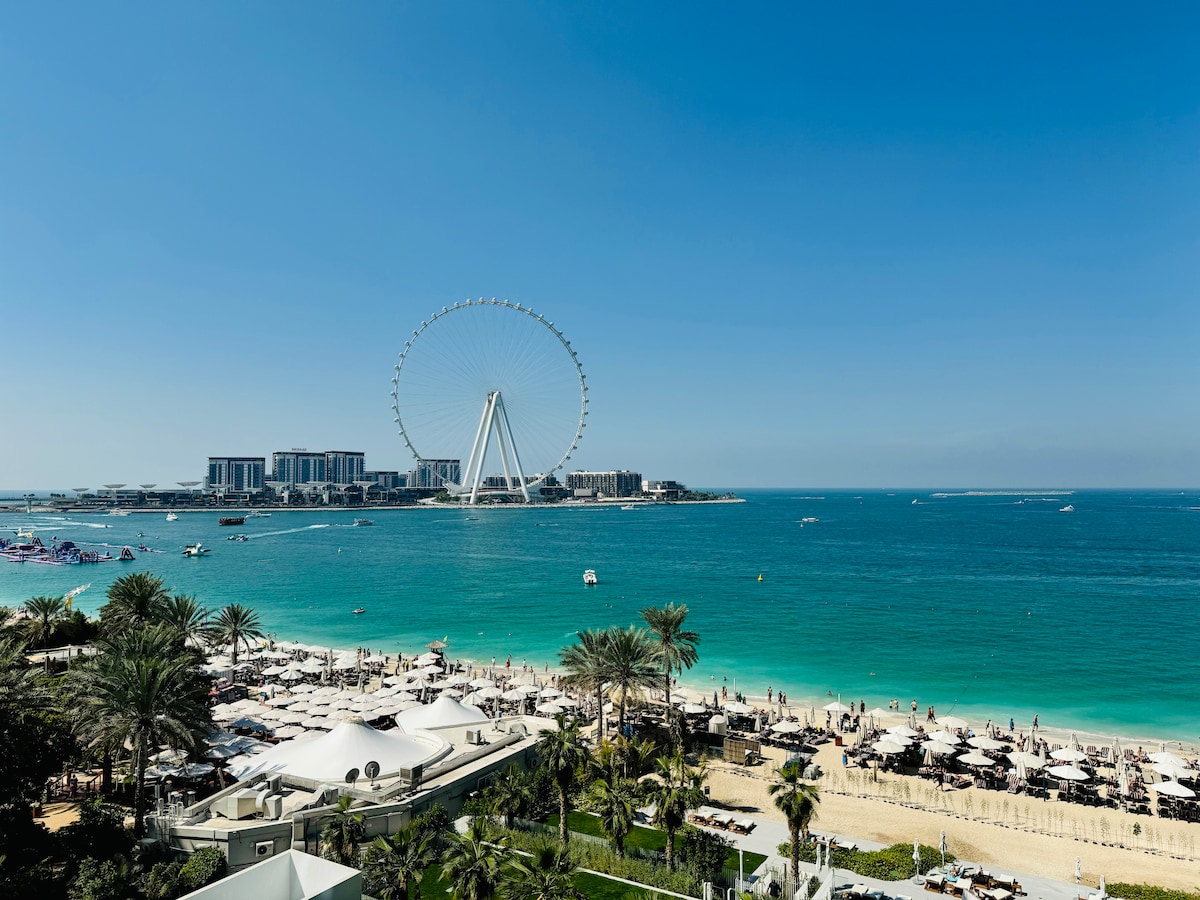 A view of the coastline is presented, showcasing a clear blue sky and the vibrant azure sea. The Jumeirah Beach area is visible along with the Dubai Eye ferris wheel in the background, complemented by sandy beaches and palm trees lining the shore.