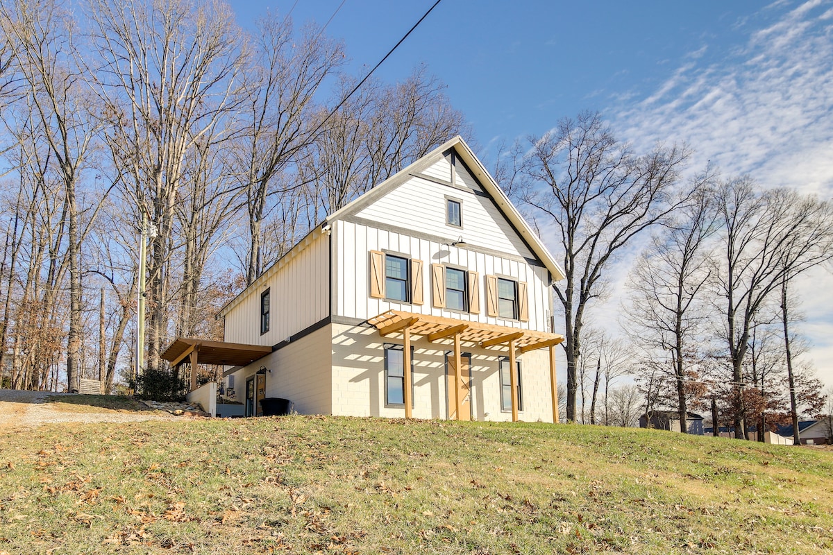 A two-story home with a modern farmhouse design is set against a backdrop of tall trees. The exterior features white horizontal siding and brown shutters, complemented by a wooden overhang at the entrance. A well-maintained lawn extends in front.