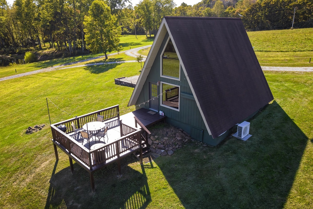 A charming A-frame cabin is surrounded by a lush green landscape, with a spacious outdoor deck featuring a table and chairs. Sunlight casts shadows on the exterior, highlighting the unique architectural design and the tranquility of the setting.