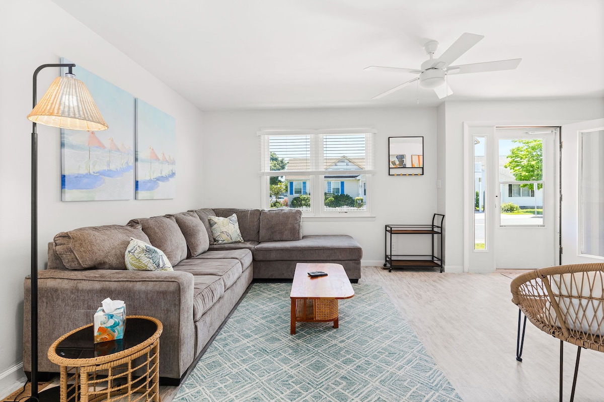 A spacious living area is furnished with a large sectional sofa and complemented by a coffee table. Natural light is invited through the windows, while a ceiling fan adds to the comfort. Decorative wall art enhances the serene ambiance.