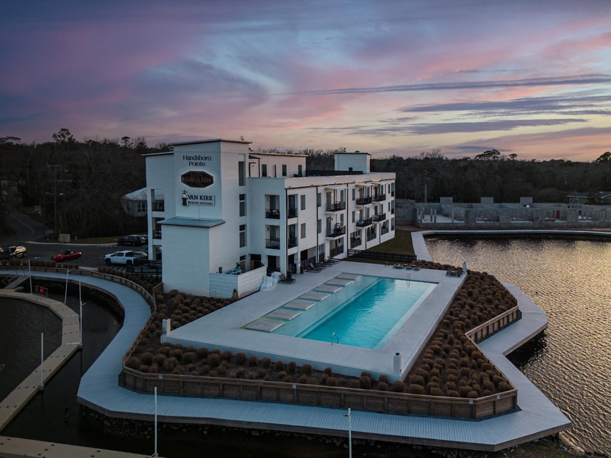 The modern multi-story building is positioned next to a reflecting pool, with an expansive outdoor deck surrounding a long, clear swimming pool. Soft hues of sunset illuminate the sky, and landscaped areas with neatly arranged shrubs frame the property.