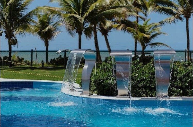 A swimming pool features three stainless steel spouts cascading water into its clear blue surface. Lush green palm trees surround the area, providing a tropical ambiance. The backdrop displays an expansive view of the ocean, enhancing the sense of tranquility.