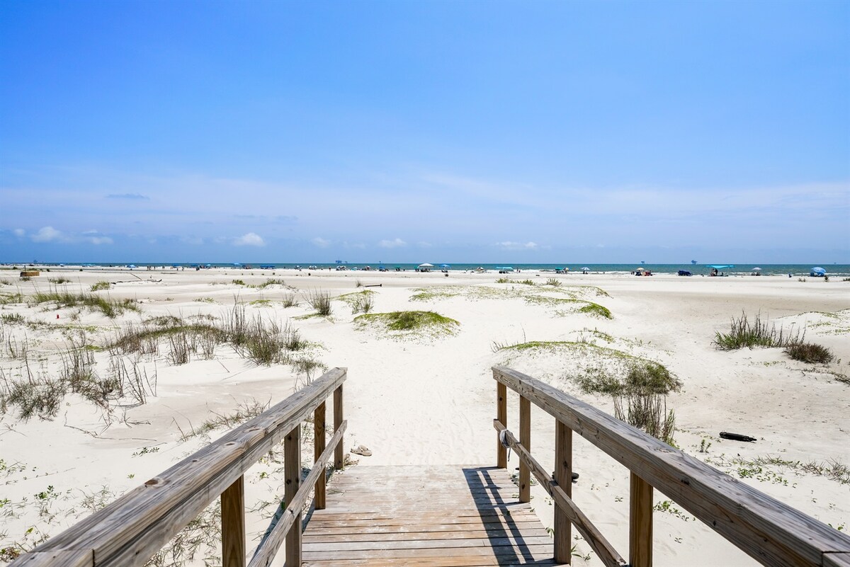 A wooden walkway leads to a sandy beach, bordered by patches of grass and gentle dunes. The expansive sea stretches in the background under a bright blue sky, with a few boats visible on the horizon.
